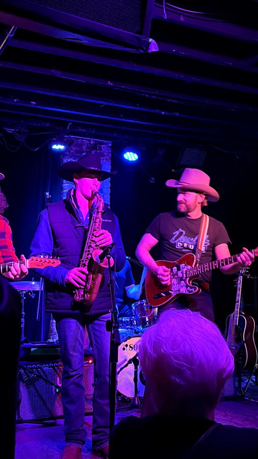 Davis in a black hat playing alto sax beside a guitarist in purple club lighting