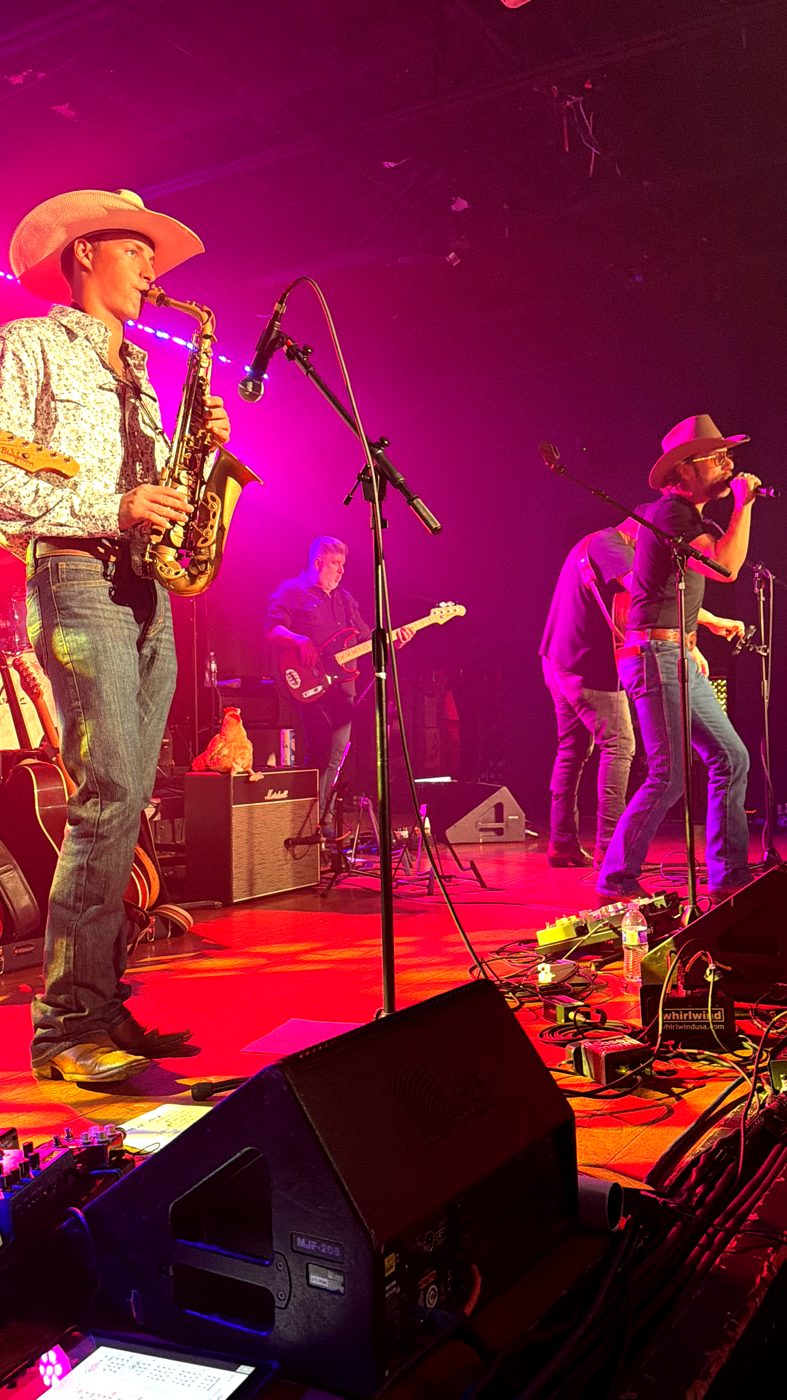 Davis Kidder in a white cowboy hat leaning into a alto sax solo, lit in pink stage light